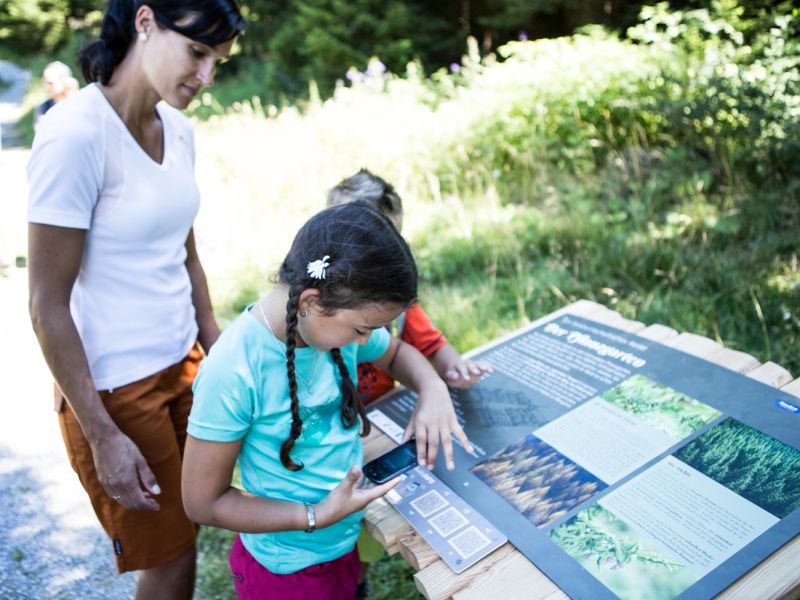 Small girl scans a code from a horizontal info board along a trail with a woman standing by.