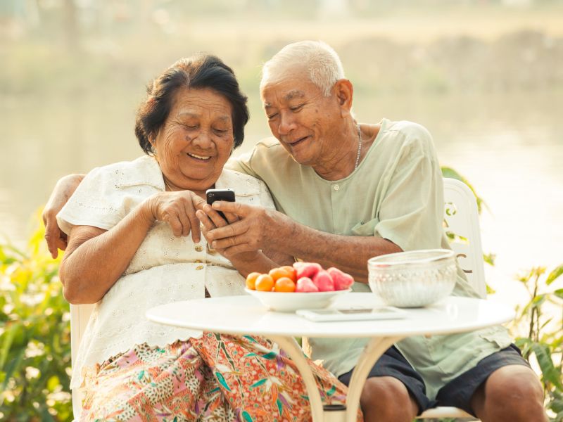 Happy senior couple sitting at a garden table, using the mobile phone
