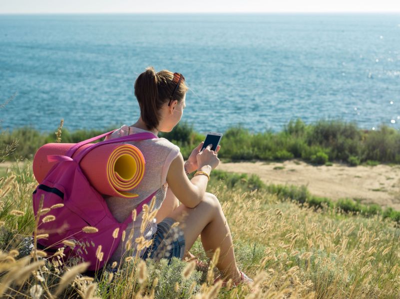 Junge Frau mit Smartphone sitzt auf einem Hügel über dem Meer