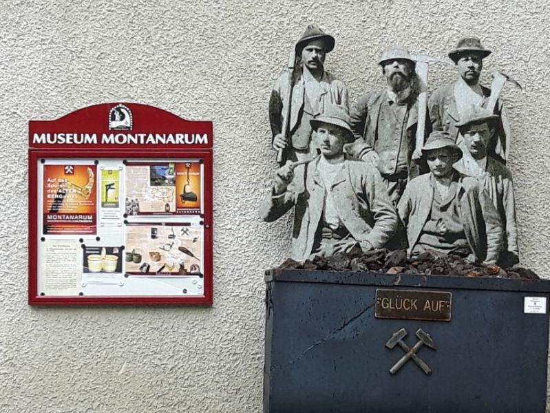 Barrierefreie naturparke - Eingang zum Montanarum in Neuberg an der Mürz mit roten Schaukasten mit der Überschrift Museum Montanarum und einen Metallkasten mit der Aufschrift "Glück auf", darüber 6 Bergmänner