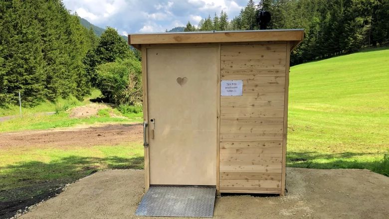 Barrierefreie Bio-Toilette mit kurzer, nur leicht geneigter Metallrampe und Schiebetür aus Holz. Im Hintergrund Wiese, Wald und Berge.