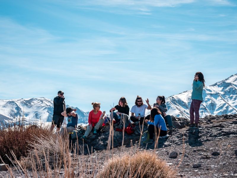 8 Personengruppe mit Wanderführer rastet in steinigem Gelände. Im Hintergrund hohe, schneebedeckte Berge